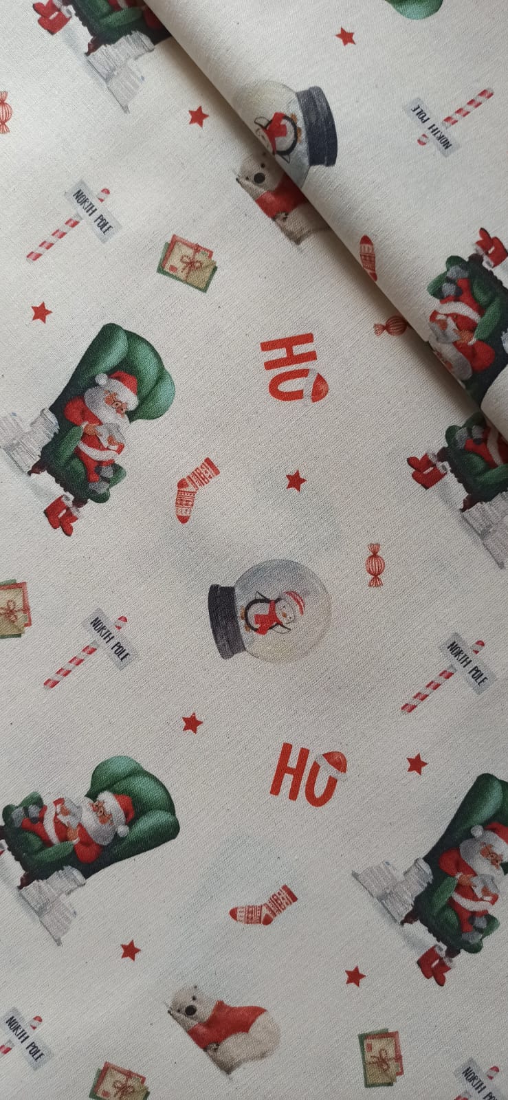 Tela de algodón con Papá Noel en sillón verde, bolas de nieve, regalos y estrellas rojas sobre fondo crema.